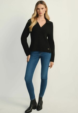 Czarny prążkowany sweter z dekoltem w serek i szerokim kołnierzem, w połączeniu z niebieskimi rurkami i czarnymi botkami za kostkę. Widoczny prosty złoty akcent.