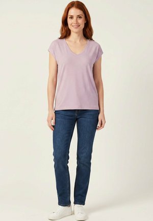 Femme souriante aux cheveux roux portant un haut violet clair à manches courtes, un jean bleu et des baskets blanches, debout devant un fond clair uni.