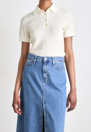 Kvinde iført en cremefarvet, ribstrikket poloshirt med korte ærmer, kombineret med en høj taljet blå denimnederdel med en slids foran.