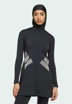 Femme portant une tunique de sport noire à manches longues avec des motifs géométriques réfléchissants et un hijab de sport noir assorti ainsi que des leggings.