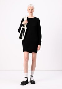 Robe en tricot côtelé noire à manches longues, associée à un sac à épaule en fausse fourrure blanche. Le mannequin porte des chaussettes blanches et des chaussures noires à semelles épaisses.