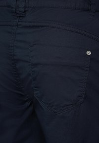 Marina blå bomullbyxor har en bakficka med en silverfärgad knappramat och synlig sömnad. Tyget har en mjuk textur.