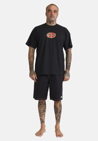 Zwarte katoenen t-shirt met een rode grafische logo, gecombineerd met zwarte shorts met witte stiksels en zijzakken. Model staat blootvoets.