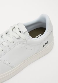 Hvit lær sneakers med teksturerte sider, flat snøring, minimale perforeringer og en beige gummisåle. Merkevarelogo trykket på siden.