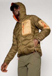 Olijf groene pufferjack met een gequiltde textuur, oranje accenten en twee borstzakken. Beschikt over een capuchon en een ritssluiting aan de voorkant.