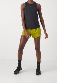 Camiseta deportiva negra sin mangas combinada con pantalones cortos verde brillante; ambos con la marca Nike. Se lleva con zapatillas deportivas negras y calcetines.