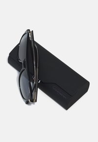 Ochelari de soare negri cu lentile întunecate, având un design elegant și accente aurii, așezați pe un toc mat negru cu inscripția "CARRERA."