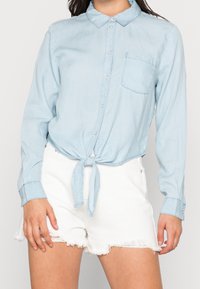 Camicia in denim azzurro chiaro con nodo frontale, chiusura a bottoni, colletto e una tasca sul petto, abbinata a pantaloncini bianchi strappati.