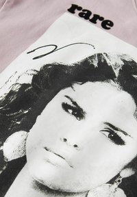 Różowa bawełniana koszulka z dużą czarno-białą grafiką twarzy kobiety, z napisem "rare" w formie pogrubionego czarnego tekstu powyżej.
