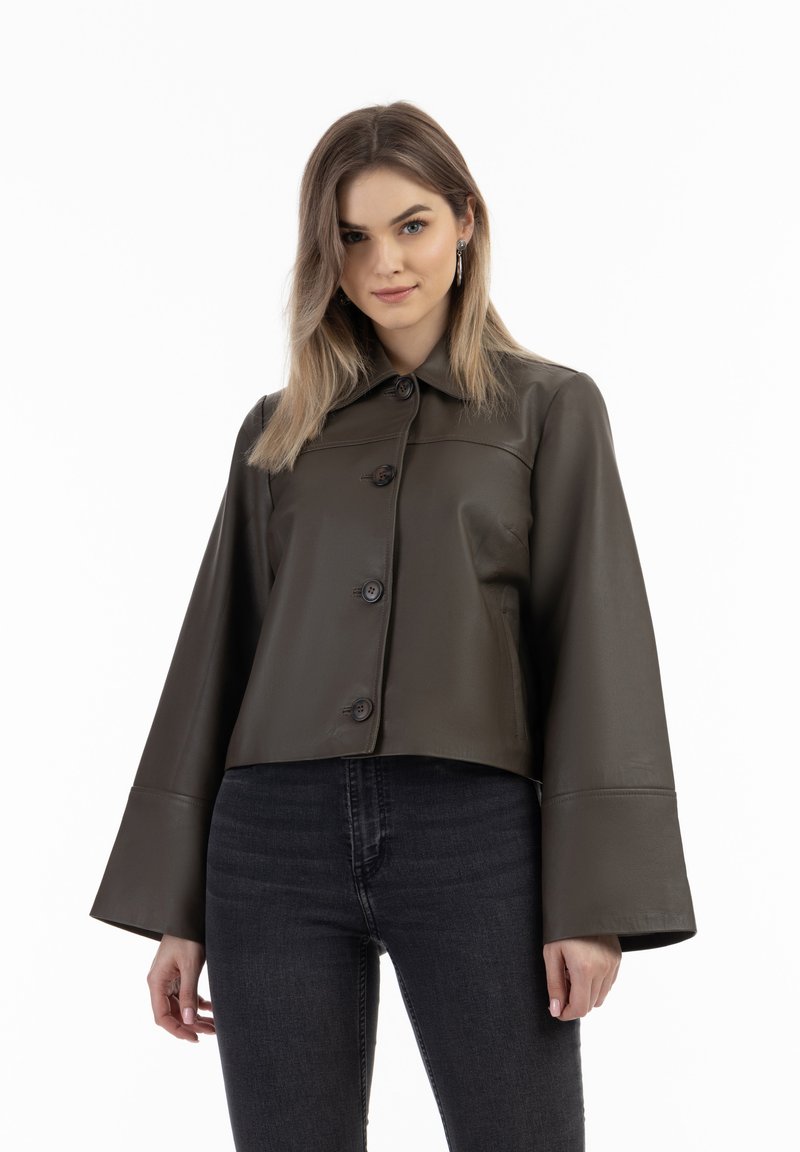 Dunkelolivgrüne Lederjacke mit Kragen, Knopfverschluss und weiten Glockenärmeln, kombiniert mit schwarzen Jeans. Glatte Textur über das gesamte Kleidungsstück.