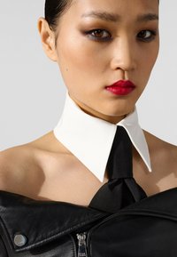 Chaqueta de cuero negra con cremallera plateada, que presenta un prominente cuello blanco y detalle de corbata negra. Textura suave, diseño moderno.