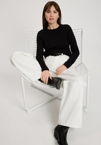 Czarna bluzka z długim rękawem w połączeniu z białymi szerokimi spodniami. Model nosi czarne botki za kostkę, siedząc na białym krześle z drutu. Prosty, nowoczesny design.