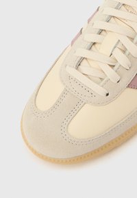 Bež velur in usnjen športni copat s kremno stransko ploščo, svetlo sivimi vezalkami in teksturirano gumijasto podplatom. Okrasen z nežnim vijoličastim detajlom.