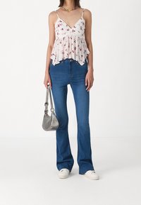 Top peplum floreale bianco con spalline regolabili, abbinato a jeans a zampa blu. Il modello tiene una borsa argentata e indossa scarpe da ginnastica bianche.