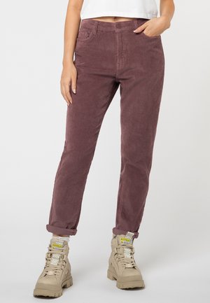 Persoon die opgerolde paarse corduroy broek en beige stevige veterlaarzen draagt, staat tegen een lichte achtergrond.