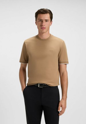 T-shirt à manches courtes, col rond, en tissu beige clair, arborant un logo discret et des finitions blanches contrastantes sur les manches. Porté avec un pantalon sombre.