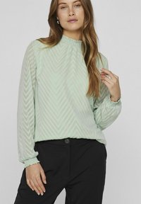 Light green blouse with a textured chevron pattern, gathered neckline, and cuffed long sleeves. Paired with black trousers.