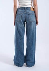 Jean en denim bleu à jambes larges avec un lavage délavé, présentant une taille haute, deux poches arrière et une étiquette de marque en cuir marron sur la ceinture.