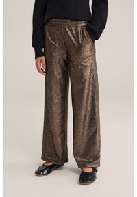 Metāliska bronzas platā kāju bikses ar teksturētu virsmu, ar elastīgu jostasvietu un sānu kabatām, kopā ar melnām patentētām plakanām kurpēm.