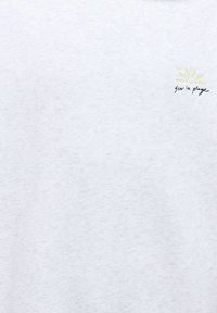 Tissu blanc avec un petit soleil jaune et le texte noir « sur la plage » brodé dans le coin supérieur droit.