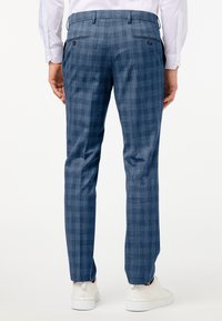 Man som bär blå rutiga byxor, vit skjorta och vita sneakers, sedd bakifrån mot en enkel vit bakgrund.