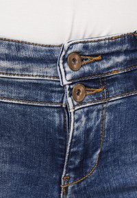 Jeans de denim de cintura alta, con dos botones de metal marrón, costuras dobles en amarillo y un lavado claro con detalles de desvanecimiento.