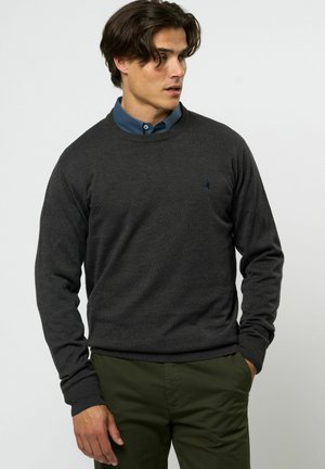 Donkergrijs gebreid sweater met een ronde hals, geribde boorden en zoom. G gedragen over een blauwe blouse met kraag, met een subtiele logodetail op de borst.