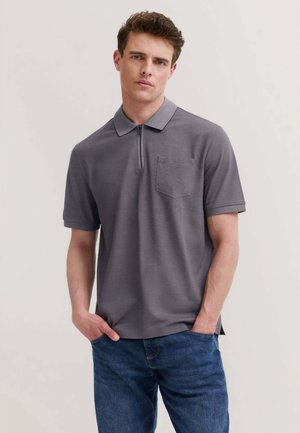 Giovane uomo con capelli castani ricci che indossa una polo grigia a maniche corte con collo con zip e jeans blu, mani nelle tasche, sfondo neutro.