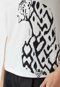 White T-shirt with a black abstract tiger print, featuring bold lines and patterns, paired with dark pants visible at the bottom edge.