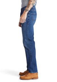 Pantalones vaqueros azules de mezclilla con un corte recto, que cuentan con un diseño clásico de cinco bolsillos. Se combinan con botas marrón claro con cordones.