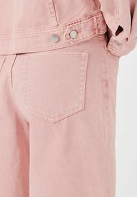 Rosa denimoverall med en litt teksturert overflate, doble metallknapper og en enkelt baklomme med rektangulær form.