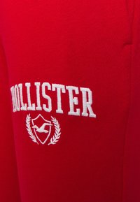 Röda sweatpants med en slät yta som har en vit broderad "Hollister"-logotyp och skölddesign med lagerbladseffekter.