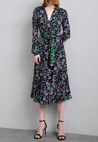 Černé zavinovací šaty s živým fialovým a zeleným květinovým vzorem, dlouhými rukávy, výstřihem do V, páskem v pase a splývavým lemem. Navrženo pro ženskou siluetu.