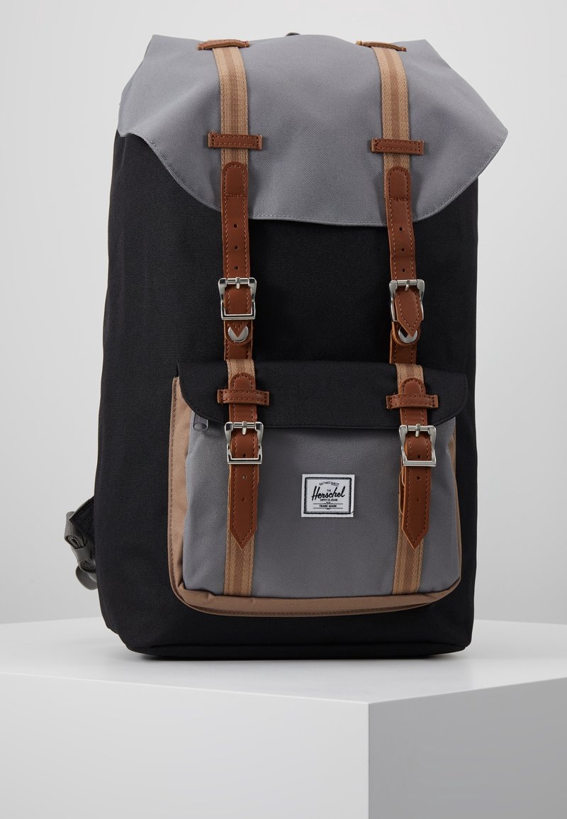 Sac à dos noir et gris avec poche avant, deux sangles à boucle couleur tan, et un écusson logo Herschel blanc sur le rabat de la poche avant.
