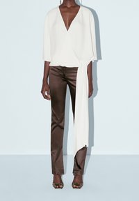 Top bianco avvolgente con maniche ampie e cravatta laterale, abbinato a pantaloni aderenti marrone scuro. Texture morbida e design elegante, indossato con tacchi aperti.