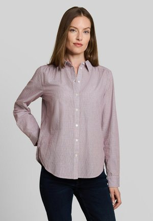 Vrouw draagt een lichtpaarse blouse met knopen en lange mouwen en een donkere broek, staand met één hand op haar heup tegen een effen achtergrond.