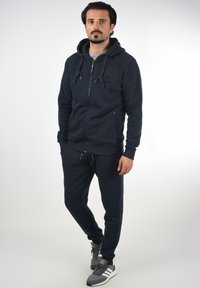 Navy blue zip-up hoodie with drawstring hood, two side pockets, ribbed cuffs, and matching pants. Sneakers with grey and white accents.