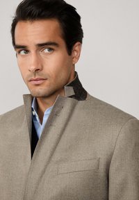 Ljust brunt blazer med en texturerad krage i mörkt material. Har en enkel ficka och ett kavajslag. Bärs över en ljusblå skjorta.