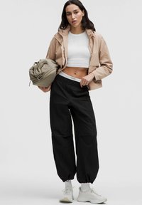 Femme portant une veste courte beige, un crop top blanc, un pantalon de jogging noir, des baskets blanches, tenant un sac de voyage beige à ses côtés.
