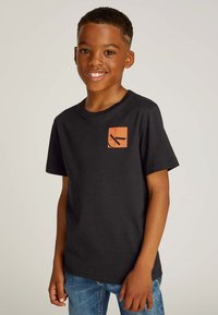 T-shirt nera in cotone con un logo quadrato arancione sul petto a sinistra. Maniche corte, scollatura tonda e vestibilità rilassata. Indossata con jeans di denim blu.