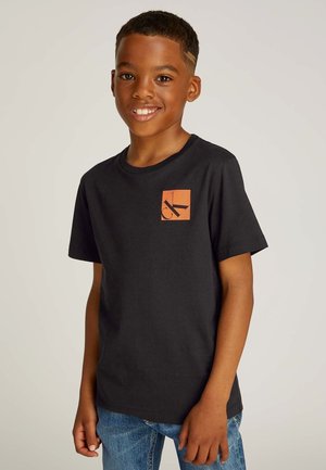 T-shirt nera in cotone con un logo quadrato arancione sul petto a sinistra. Maniche corte, scollatura tonda e vestibilità rilassata. Indossata con jeans di denim blu.