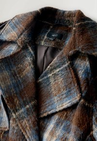 Brun og blå plaid frakke lavet af fuzzy stof, med en stor krave, forlommer og en glat brun indvendig foring. Mærkets label er synlig.