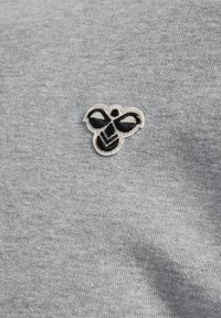 Grå bomullstyg med en liten broderad bi-logotyp i svart och vitt, placerad på vänster sida. Ytan verkar slät och enhetlig.