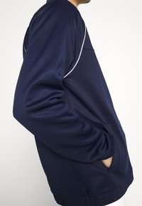 Navy athletic jacket with a textured finish, featuring white piping along the shoulders and a kangaroo pocket. Relaxed fit with elastic cuffs.