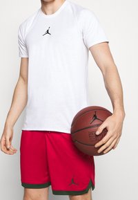 Persona vistiendo una camiseta blanca de Jordan y pantalones cortos rojos, sosteniendo un balón de baloncesto marrón con el logo de Jordan, frente a un fondo liso.