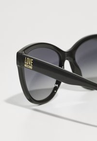 Óculos de sol pretos com um acabamento brilhante, apresentando lentes escuras em gradiente. Logótipo dourado "LOVE MOSCHINO" na lateral, com formato arredondado de gato-olho.