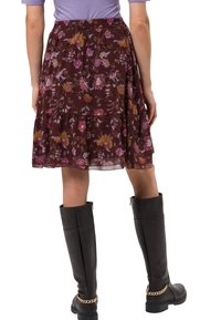 Femme portant une jupe fleurie marron à volants, des bottes noires montantes avec des chaînes dorées, et un haut à manches courtes violet clair, vue de dos.