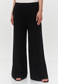 Czarne spodnie o szerokich nogawkach, wykonane z miękkiego materiału, z gładkim paskiem w talii i minimalistycznym designem, z widocznymi butami na odkrytych palcach.