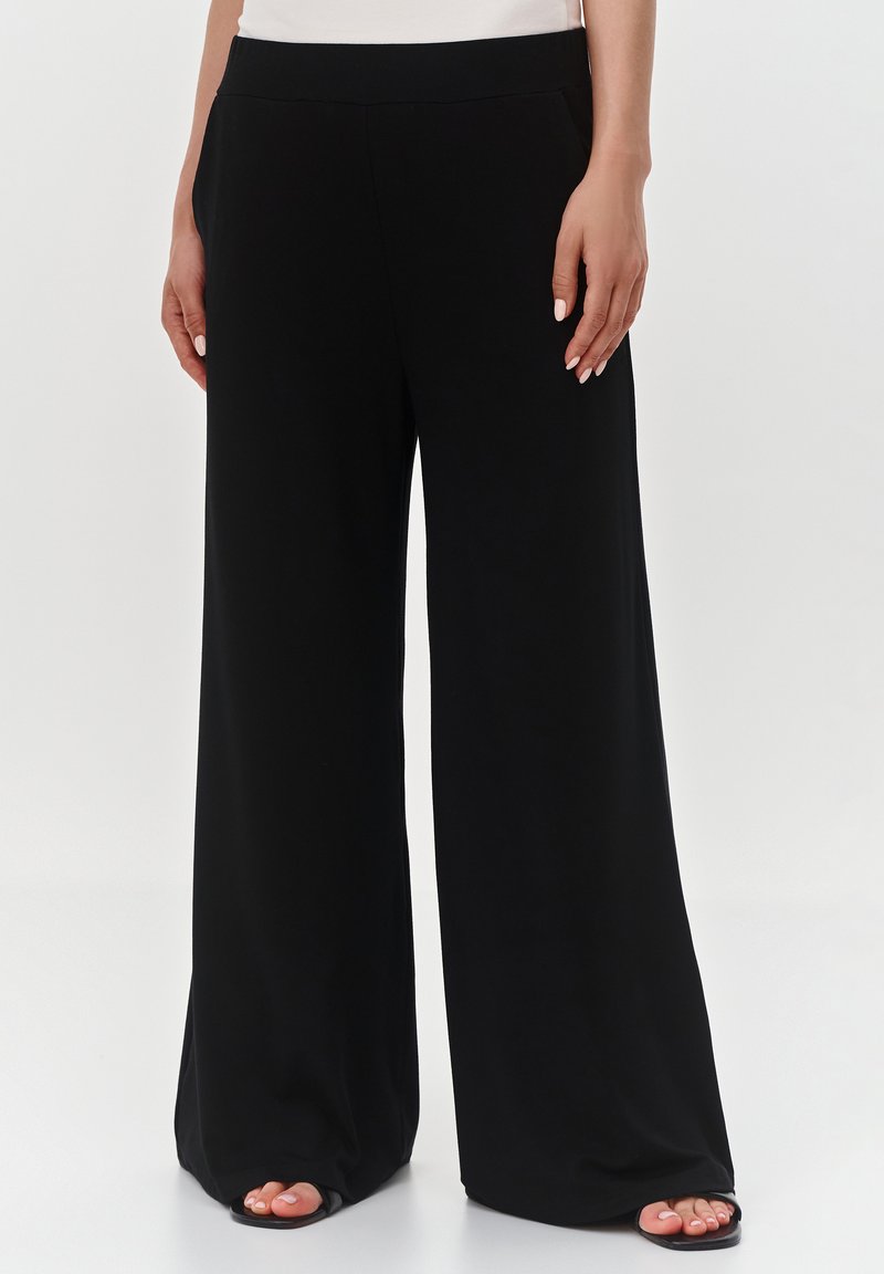 Czarne spodnie o szerokich nogawkach, wykonane z miękkiego materiału, z gładkim paskiem w talii i minimalistycznym designem, z widocznymi butami na odkrytych palcach.