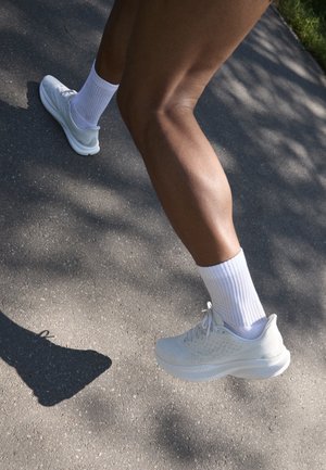 Sapatilha desportiva branca com parte superior texturada, atacadores e sola almofadada, acompanhada de meias brancas até ao tornozelo, sobre um fundo de pavimento cinza.